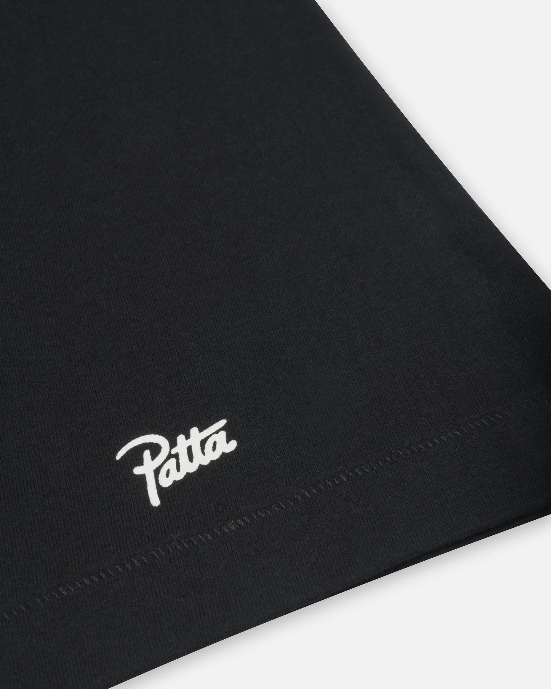 Close-up of the white Patta Script logo printed above the bottom hem on a black cotton t-shirt.